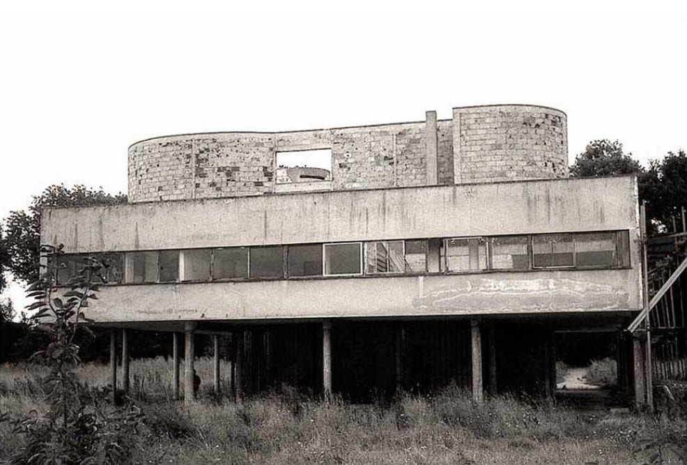 Black and white photo of an abandoned modernist building on pilotis with deteriorating brick upper level and strip windows.