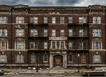 Abandoned brick apartment building with fire escapes, weathered facade, and a vintage car parked outside under overcast sk...
